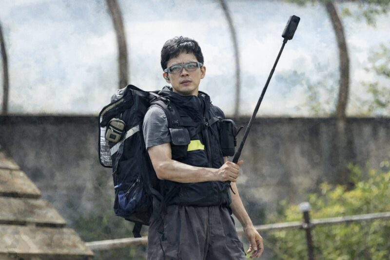 本心(2024)|リアルアバターとして装備を背負いマイクを持つ主人公の緊張感ある屋外シーン(水彩画風・フォトリアル寄り)